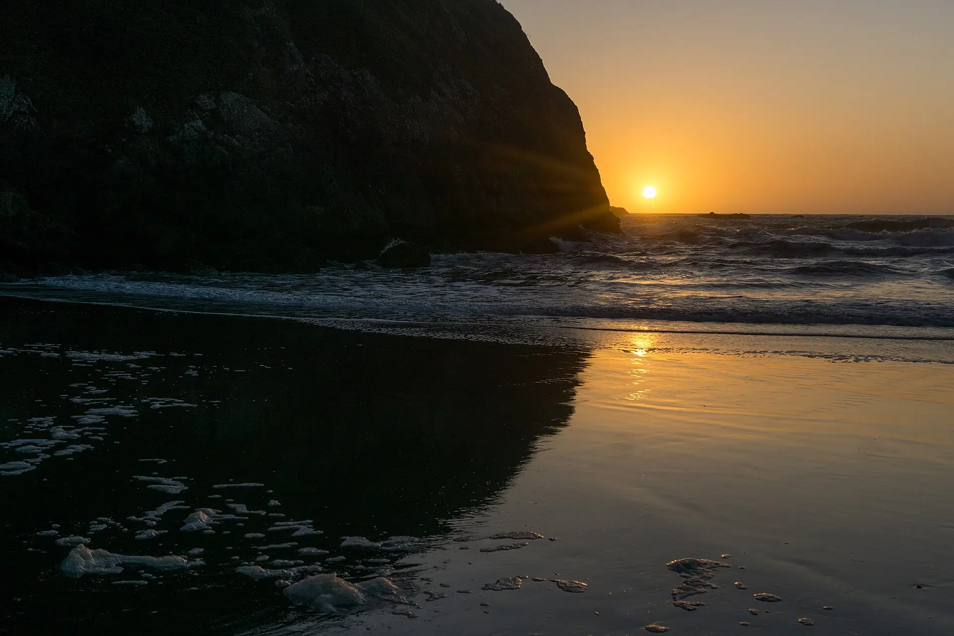 Sunset at Trinidad State Beach, the sun dropping behind a dark headland with surf reflecting gold